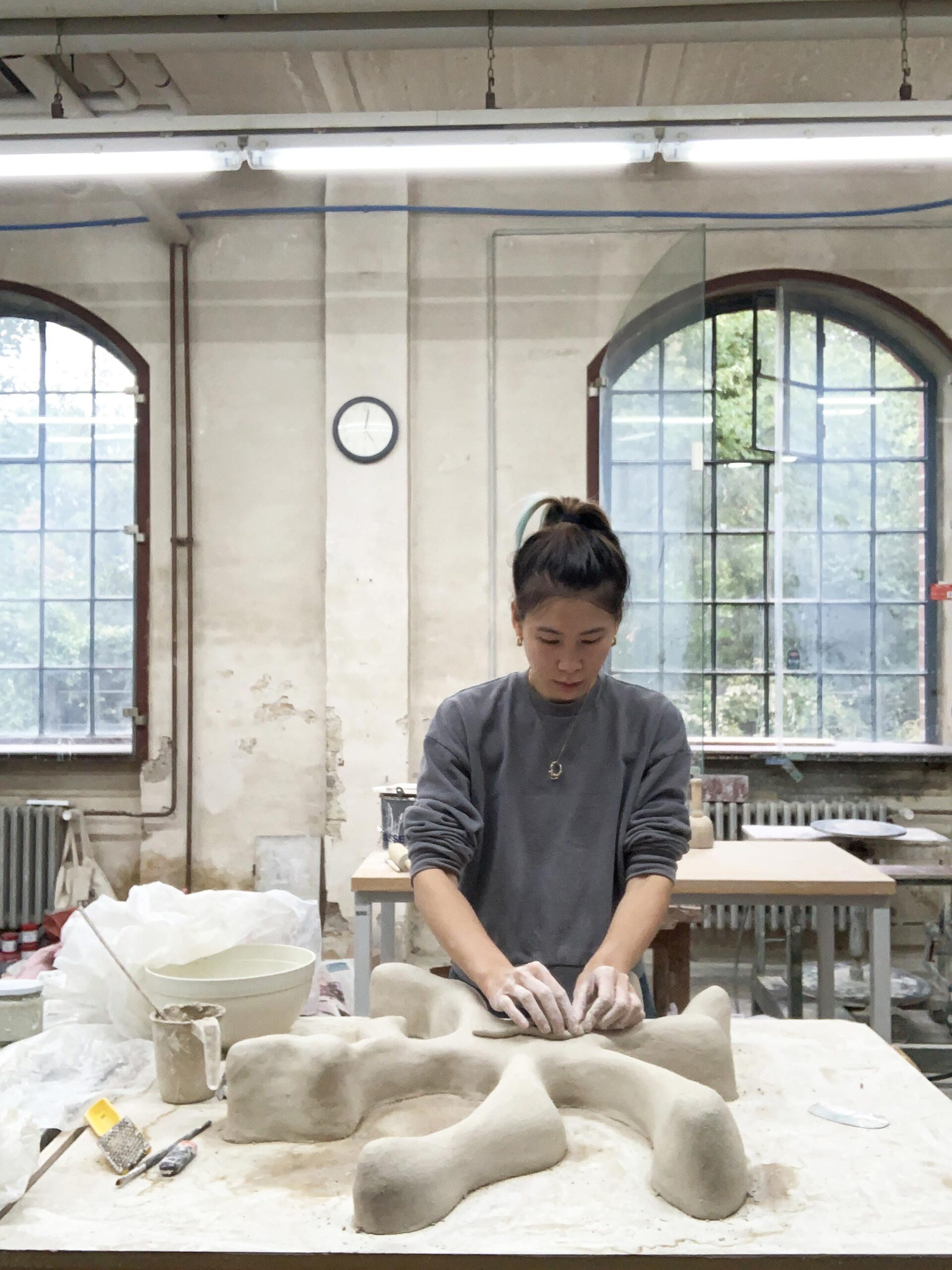 A young woman of Asian decent with dark pinned up hair, wearing a grey jumper and ring as a necklace is standing in a well-lit artist studio space close to a table working on a large light grey clay object with multiple tentacles, her hands are covered in clay mud 